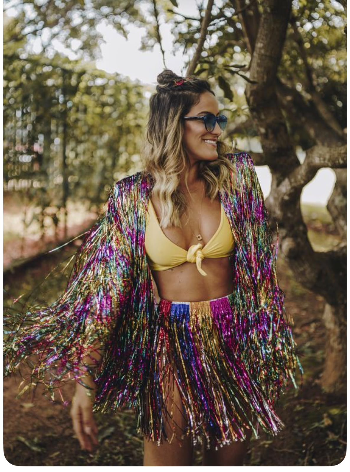 Mujer con chaqueta y falda de flecos metálicos arcoíris sobre un bikini amarillo, con gafas de corazón